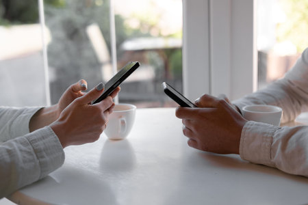 close-up of smart phones in the hands of woman and man sitting on wooden table at coffee shop....