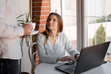 Image of smiling woman 20s working on laptop and man with coffee for wife, together while sitting at homeの写真素材