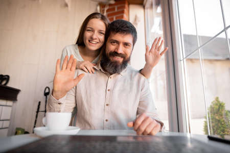Happy young european couple resting at living room, waving hand hello using webcam laptop, greeting friends at online video call distant meetingの写真素材