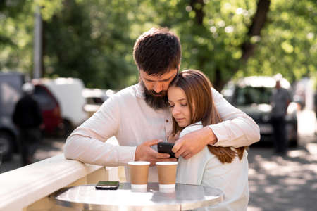 Couple in love drinking coffee in a street cafe in summer, man hugging woman and using a smartphone to call or search the internetの写真素材