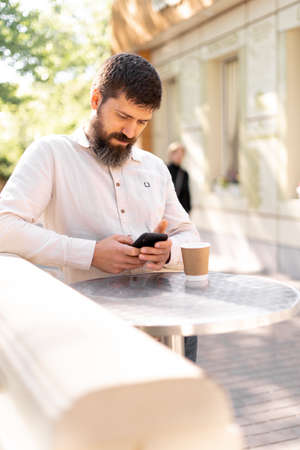Young bearded man in business shirt attire standing in relaxing outdoor cafe drinking coffee while using mobile phoneの写真素材