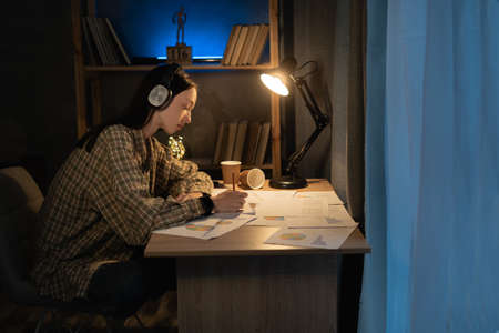 asian female freelancer working at night analyzing information in documents while sitting at table with graphics in cozy contemporary apartment, girl student work with new projectの写真素材