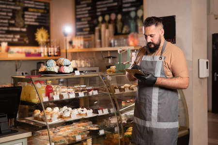 Successful small business owner in cafe using digital tablet and looking at screen. Serious barista with a beard standing in front of showcase while counting, Start upの写真素材