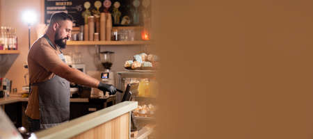 Bearded waiter in apron at counter with cashbox working at bar or coffee shop, male barista at counter in cafe store checking clients order, small business, people and service conceptの写真素材