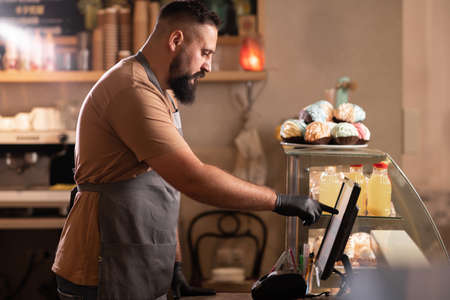 Profile view male barista at counter using cashbox computer in cafe store. Indian Waiter in apron working in coffee shop.small business, people and serviceの写真素材