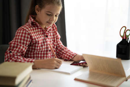 Close up focused little girl using phone studying online, sitting at table at home, serious child holding smartphone, watching webinar, writing notes, homeschoolingの写真素材