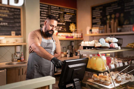 Male Indian barista or owner at counter using cashbox computer in cafe store checking clients order. Waiter in apron working in coffee shop. small business, people and serviceの写真素材