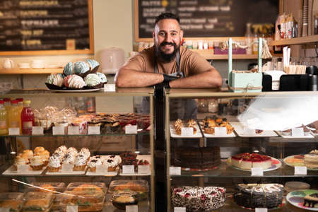 Cheerful indian male baker welcoming you at his bakery store, bearded man baker smiling to the camera proudly leaning on the showcase at his cafeの写真素材