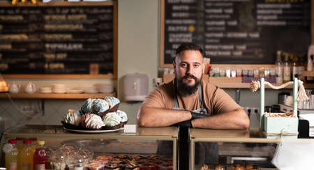 Cheerful indian male baker welcoming you at his bakery store, bearded male baker smiling to the camera proudly leaning on the showcase at his cafeの写真素材