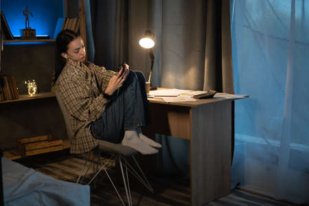 Asian teenage girl listening to the music using a cell phone and headphones at home at late night, student relaxing from study while sitting at a desk , technology, people and educationの写真素材