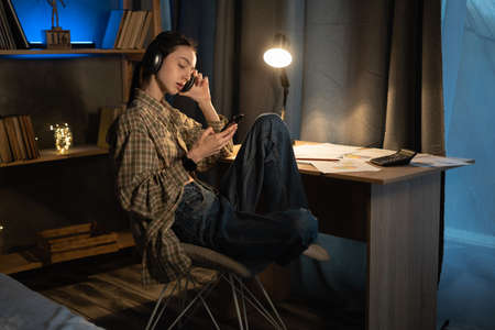 Asian teenage girl listening to the music using a cell phone and headphones at home at late night, student relaxing from study while sitting at a desk, technology, people and educationの写真素材