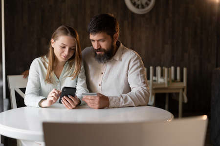 Young couple doing online shopping in cafe using smartphone, bearded man and girl chatting online while sitting in restaurantの写真素材
