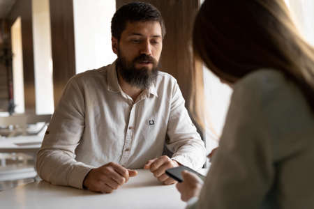 Portrait of a young bearded man at a table in a cafe looking at a smartphone with a girl sitting next.の写真素材