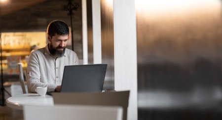 European young male in white shirt using laptop and drinking coffee in coffee shop cafe while working online freelance businessの写真素材