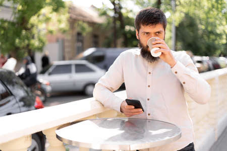 man drinking takeaway coffee standing on the street using mobile phone, bearded guy chatting on social network in street cafeの写真素材