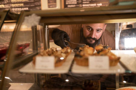 Indian man choosing variation of delicious cake in bakery showcase fridge at cafe. Male customer buying sweet cake at coffee shop. Small business restaurant foodの写真素材