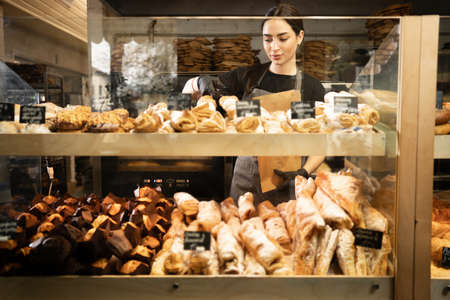 Young woman preparing pastry for sale in supermarket bakery department. Bakery seller.の写真素材