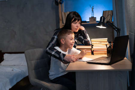 mom helping son with homework while sitting at desk at home using laptop and books at night. online schoolingの写真素材