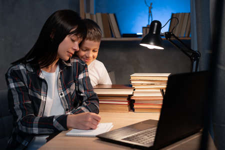 Busy mom works at home with son at night. Mother is watching on laptop while working remotely and writing notesの写真素材