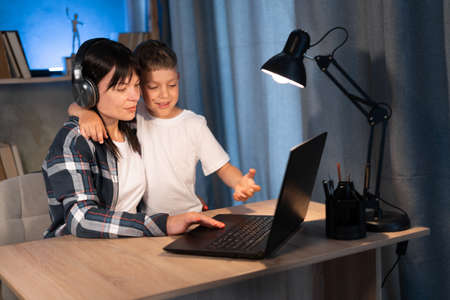 Happy young mother talking to son while working on laptop at home. boy looking for mom while typing on computer at night. woman and child while using laptop.の写真素材