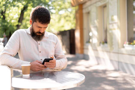 Serious male journalist having rest while drinking coffee while texting with friends on mobile phone at city streetの写真素材