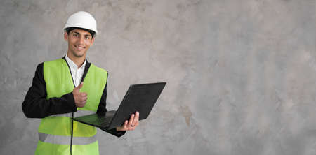 Arabic civil engineer using laptop. Engineering and construction concept. Happy engineer with laptop computer. Man in hard hat. Architect in yellow vest on grey background. Bannerの写真素材