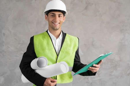 Confident young arabic male construction engineer in hardhat and waistcoat holding rolled blueprint and looking at camera with smile against grey backgroundの写真素材