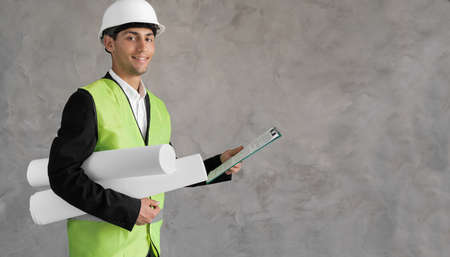 Young arabic civil engineer holding paperwork blueprint roll standing post in studio. Content male construction architect in suit and hardhat with rolled blueprints on isolated grey background.の写真素材