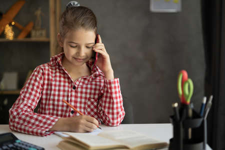 Caucasian child schoolgirl talking on the phone while studying remotely, taking notes while studying at home. chatting with friends using smartphone. E-learning. Homeschoolingの写真素材