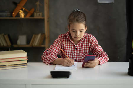 young girl doing homework at home using mobile phone, homeschooling and e-learningの写真素材