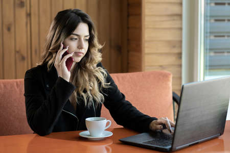 Young successful businesswoman calling with cell telephone while sitting in coffee shop, female entrepreneur using laptop and having talk with mobile phone in cafe, distant work and freelanceの写真素材