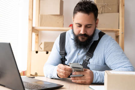 A young Indian male entrepreneur at home works on a laptop computer and online checks orders, preparing to pack boxes, sell to customers, count money dollar business delivery, smeの写真素材