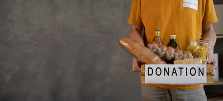 Volunteer with donation box with food, close up. A man holding a donations box of different products on grey background, bannerの写真素材