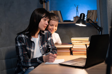 Busy mom works at home with son at night. Mother is watching on laptop while working remotely and writing notes at the table. Education technologyの写真素材