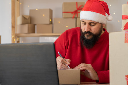 Indian bearded man wearing a Santa Claus hat works in a warehouse of boxes with gifts and orders from an online store for Christmas. Small Business Owner Sells New Yearの写真素材