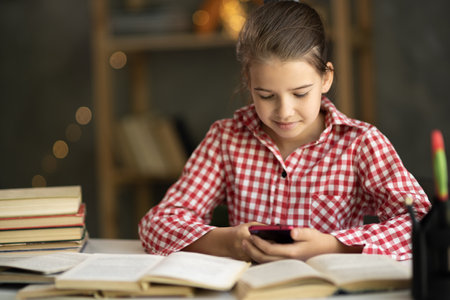 Teenage girl school pupil holding smart phone, studying with book. Focused teen student doing homework using mobile app for remote education at home. e-learningの写真素材