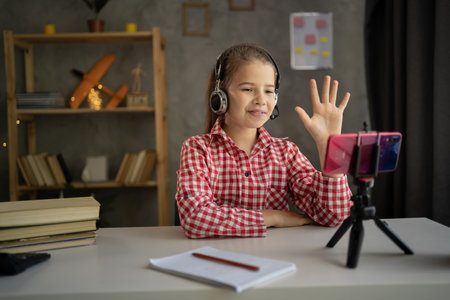 teenage girl wears headphones with online learning by phone in mobile app. college student using smartphone on tripod watching video course, zooming, sitting at home on lessonの写真素材
