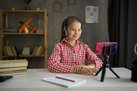 Smiling little girl wearing headphones studying online at home, looking at phone screen, engaged in video conference, watching webinar, using smartphone on tripod, homeschoolingの写真素材