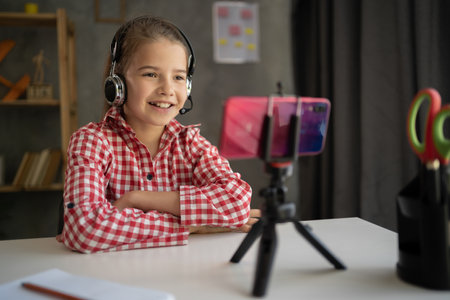 Happy student girl wearing headphones studying online at home, looking at phone screen, engaged in video conference, watching webinar, using smartphone on tripod, homeschoolingの写真素材