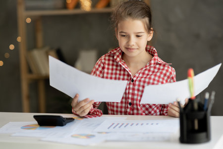 Happy little girl sitting at table reading good news in paper letter, checking internal accounts, smiling child holding paperwork, doing paperwork or studying sitting at home deskの写真素材