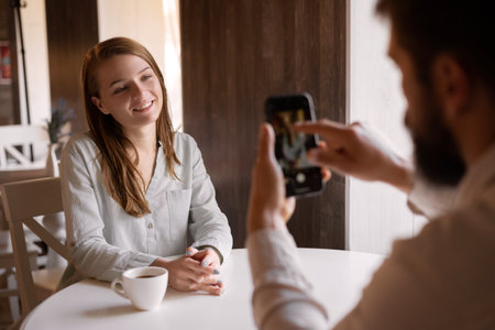 couple in love sitting in cafe, male photographer photographing woman on smartphone, indoor mobile photography, looks lovingly at boyfriend or partner, smiles. Social media blog introductionの写真素材