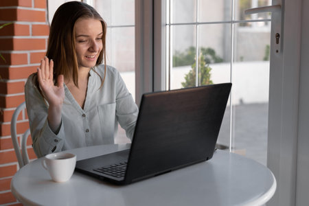 Smiling young woman using laptop and making video call in cafe. girl talking and waving hello during an online call using a laptop while sittingの写真素材