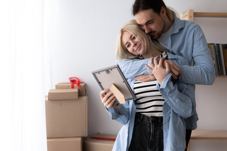 Happy wife and husband looking at photo frame, sharing memories, standing at new home, young couple unpacking cardboard boxes with belongings on moving day, relocation and mortgageの写真素材
