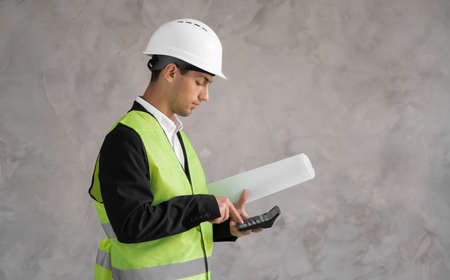 The cost of a new home. Muslim young man with drawings and a calculator isolated on grey background. Estimate of construction. A Builder in a white helmetの写真素材