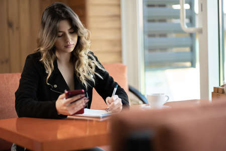 Intelligent female student writing text to education notebook sitting at cafe table and using smartphone, young girl with mobile phone making notesの写真素材