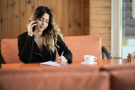 Young business woman sitting at table in cafe, talking on cell phone while taking notes in notebook. Student learning online. Freelancer workingの写真素材