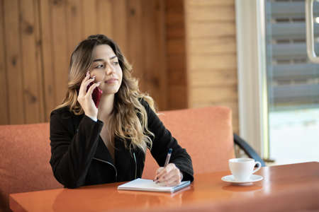 Young beautiful girl makes a phone call in a cafe, writes notes in a notebook. Student learning online. Freelancer workingの写真素材