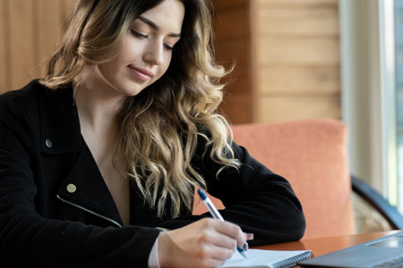 Portrait of a young business woman sitting at a cafe table and making notes in a notebook using a laptop. Teaching students online.の写真素材