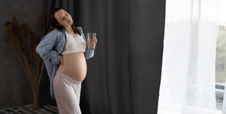 Pregnancy backache. Pregnant woman having lower back pain standing near window and drink clear water. Pregnancy ache and problem concept. bannerの写真素材