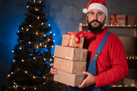 Small and Medium Business Startup Concept A young start-up businessman, wearing a santa claus hat and work overalls, takes a stand in a home office holding a lot of boxes in his hands.の写真素材